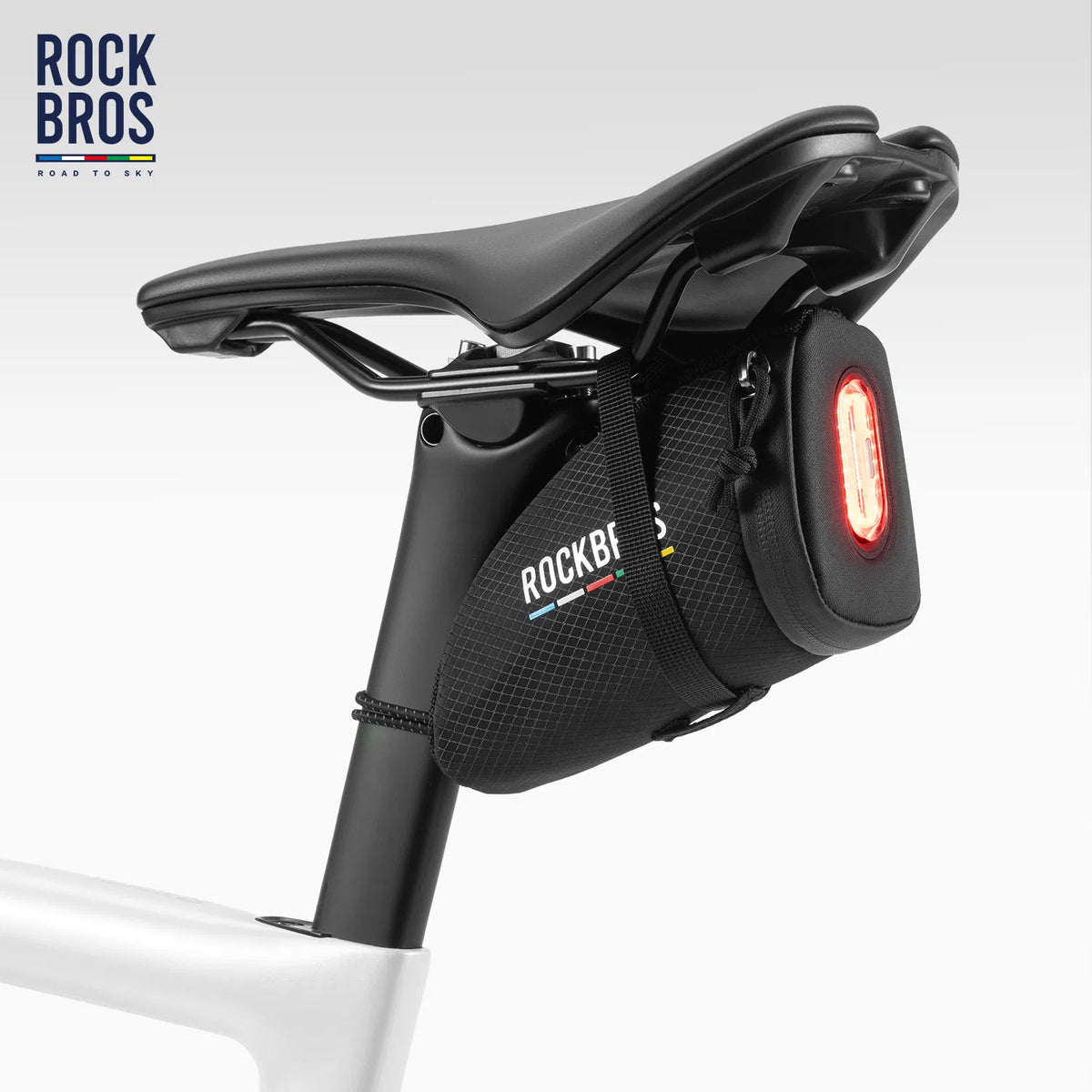 A black ROCKBROS rear rack bag mounted on a bicycle seat post, featuring a red rear light and white brand logo, shown against a light gray studio background.