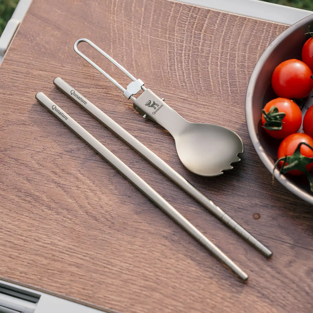 Set of silver utensils including a spoon and two straws on a wooden surface with tomatoes in the background.