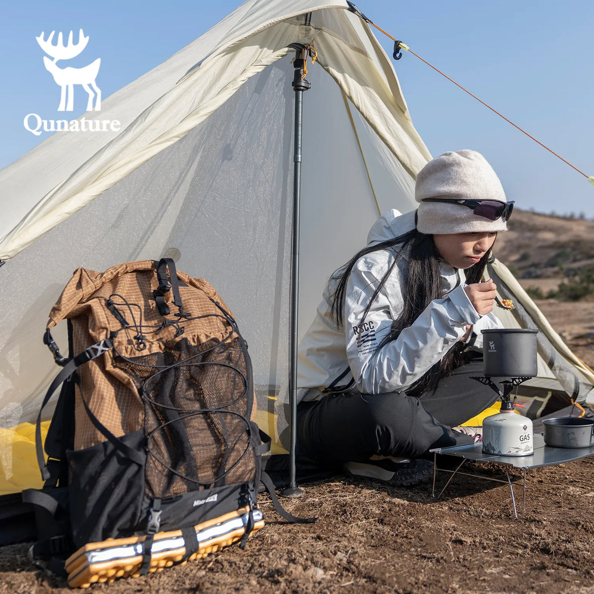 A khaki hiking backpack was placed on the ground, and a girl was sitting in a tent next to it eating.