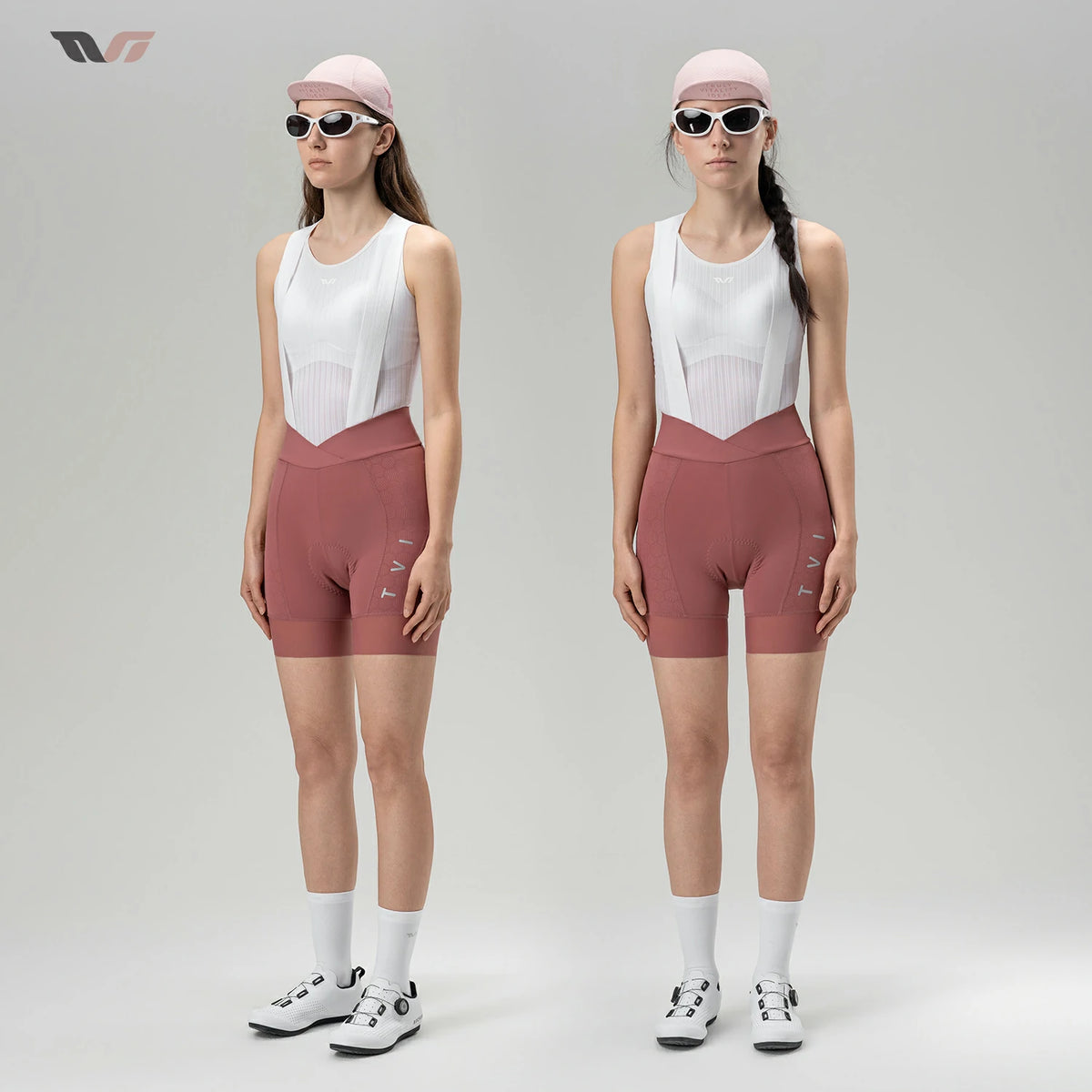 Two female models standing side by side in a studio, wearing matching athletic cycling outfits consisting of white bib shorts and dusty rose fitted shorts, white socks, and cycling shoes. Both wear light pink caps and dark sunglasses against a light gray background.
